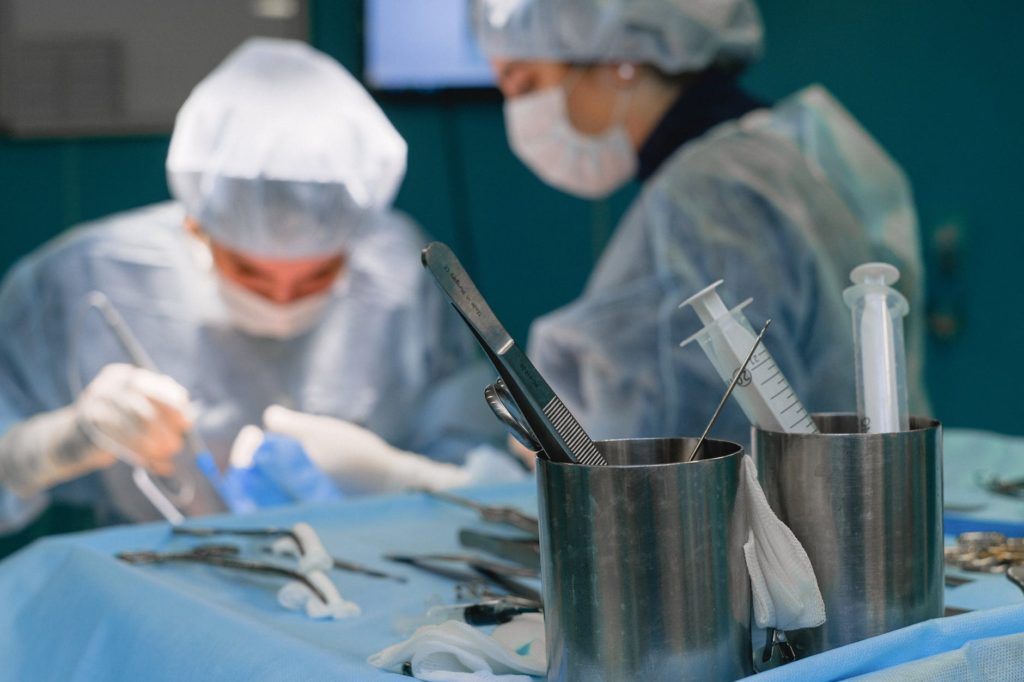 two surgeons in the operating room wearing masks, caps, gowns, and gloves, performing an operation; a container and tray of surgical instruments is shown in front