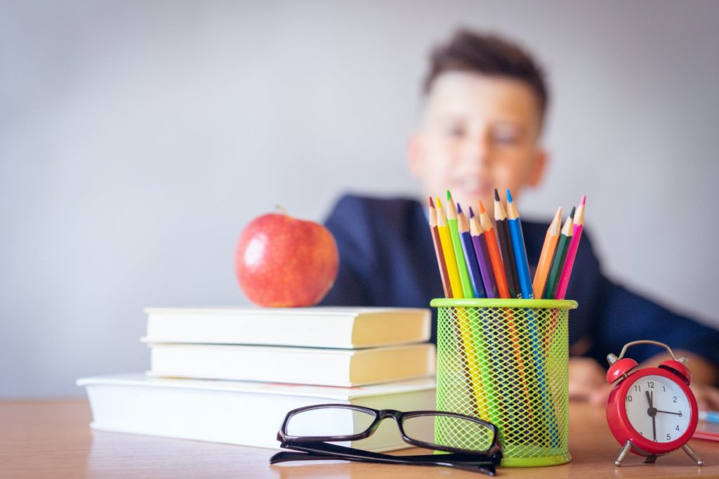 Boy sitting behind a desk. On the desk there is a pair of glasses, a cup of pencil crayons, a small clock, a pile of books, and an apple atop of the books.