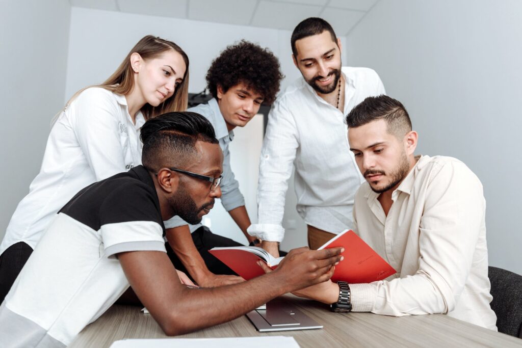 a multi-ethnic group of adults gathered around a table looking at an open book together