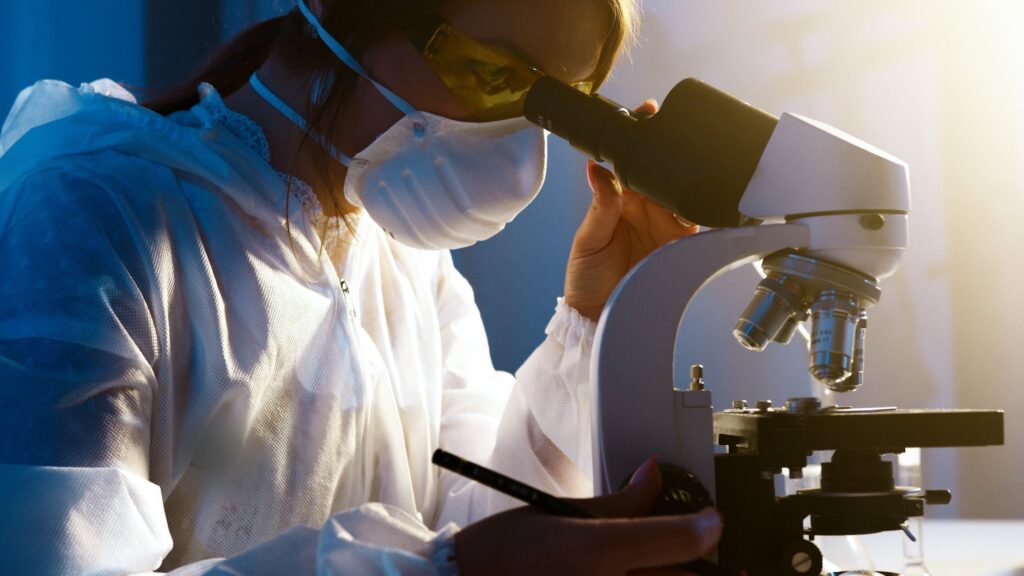 a person wearing a mask and white gown looking into a microscope