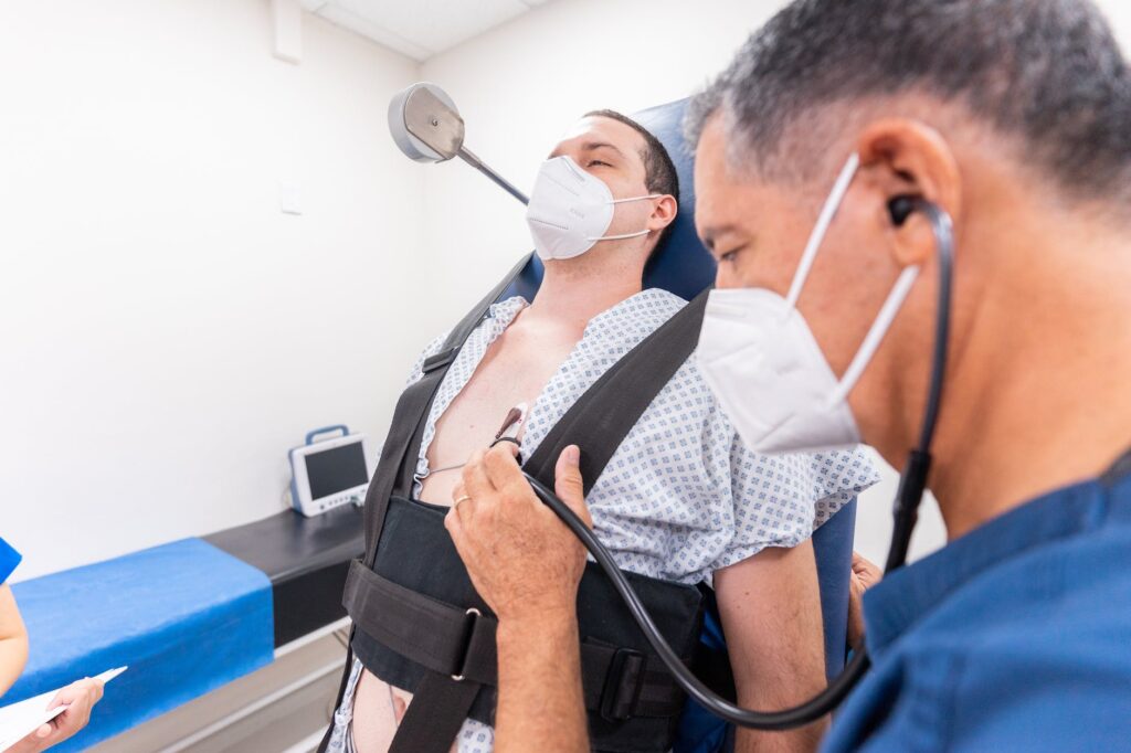 a healthcare professional and a patient, both wearing masks. The healthcare professional is listening with the earpieces of the stethoscope in his ears while the bell of the stethoscope is applied to the patient's chest