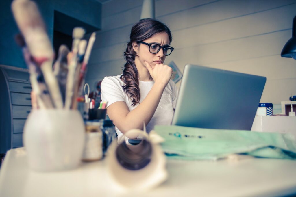 alt="a woman wearing glasses seated at a desk looking at her laptop."