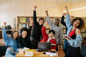 a group of students behind a desk at a library gathered for a group photo smiling with their fists pumped into the air