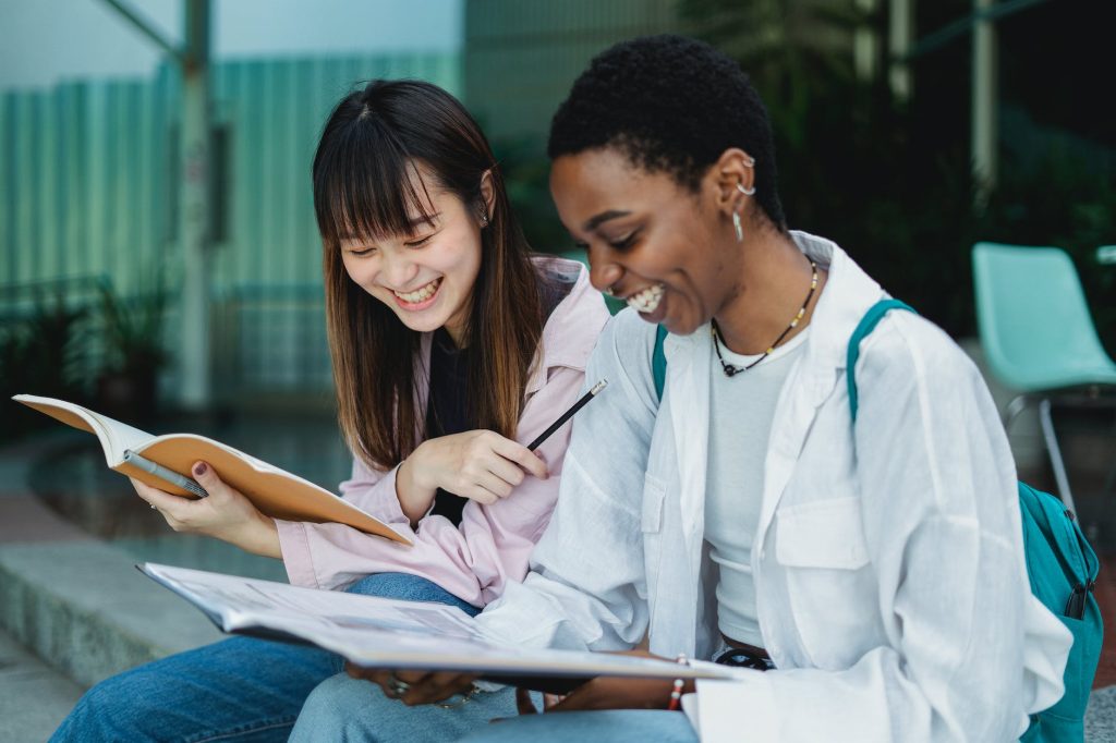 Two students seated together and smiling as they are looking down at the books that they are holding open