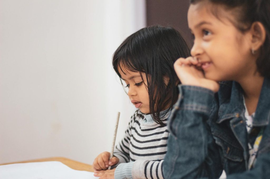 two children seated at a school desk, one is looking down and writing with a pencil and the other is looking ahead and smiling