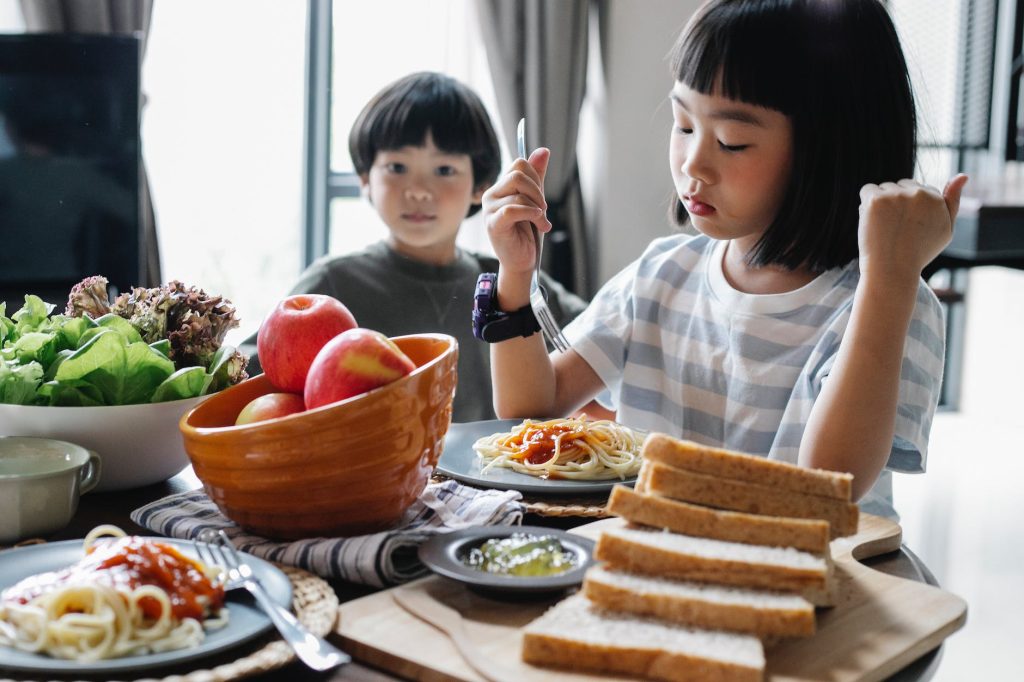 two children sitting at a table for a meal with bread slices and a bowl of apples in front of them