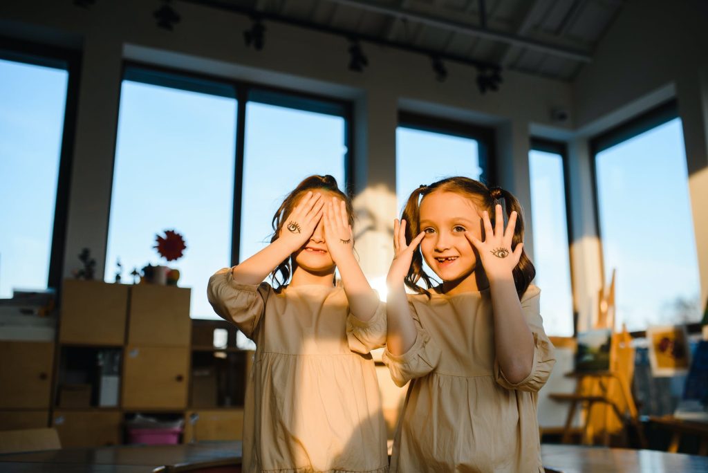 two children looking ahead and smiling, seeming to play peek-a-boo, one of them is covering her eyes, and the other one has her hands by the side of her face