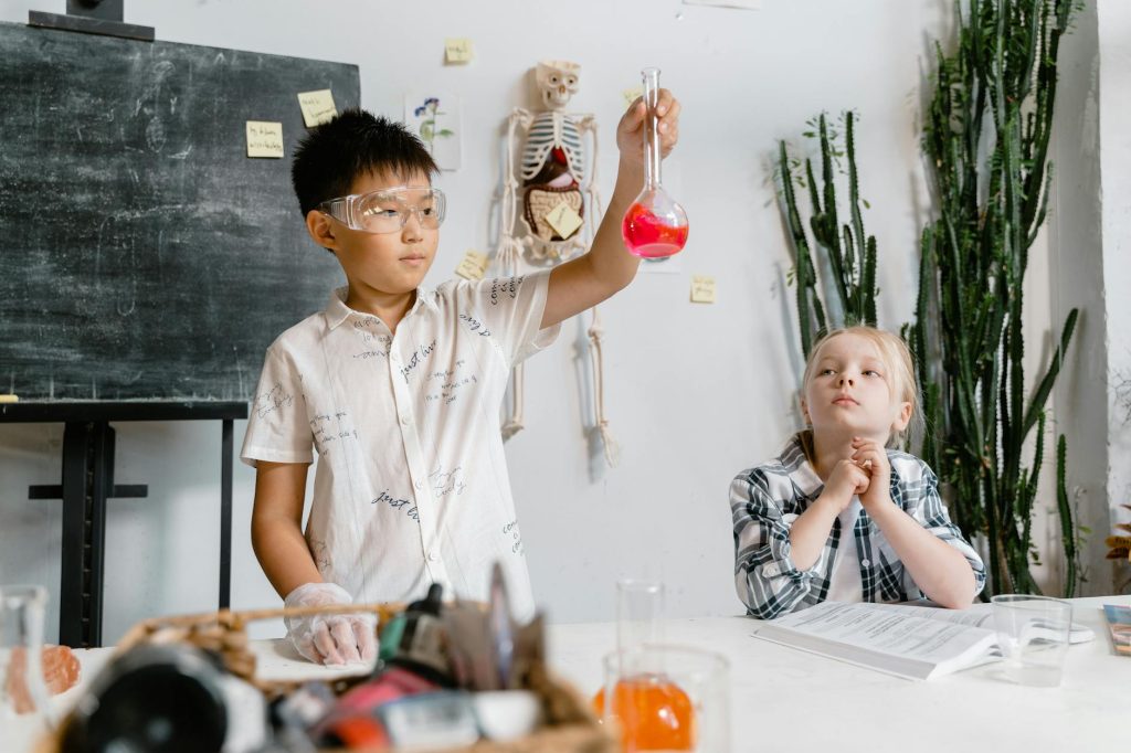 two kids at a table, one is sitting and the other one is standing while wearing safety glasses and holding a beaker containing red liquid