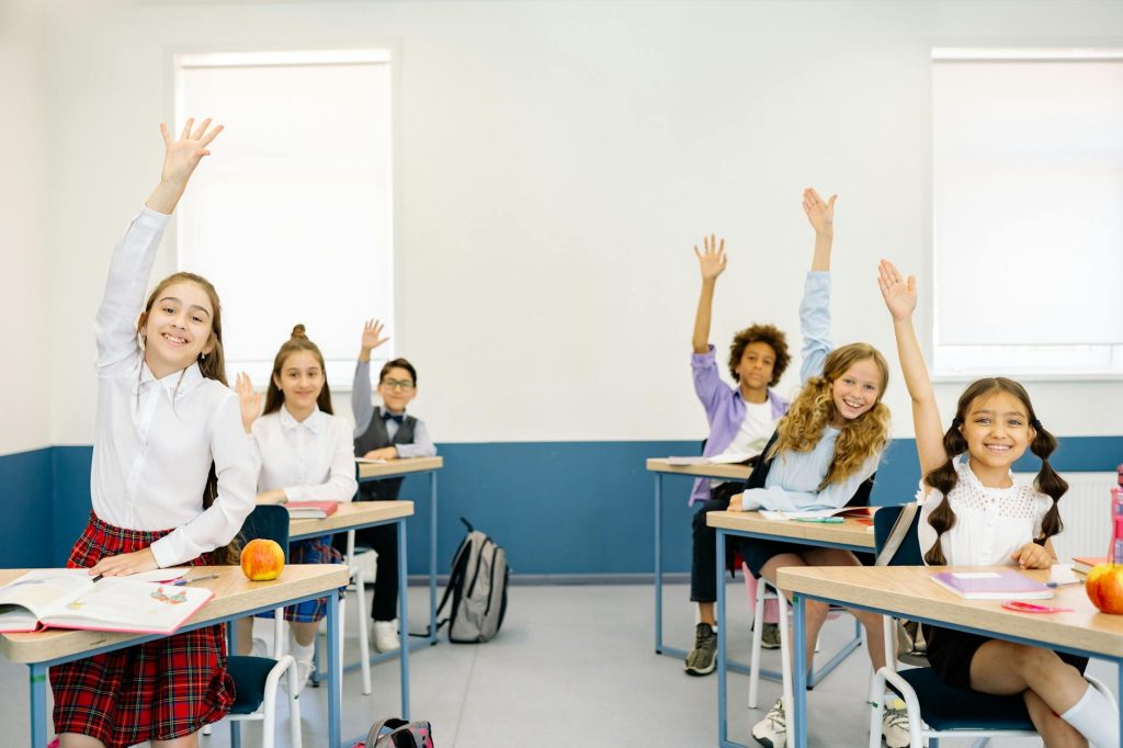 children in a classroom and all of them are raising their hands while they are seated at their desks