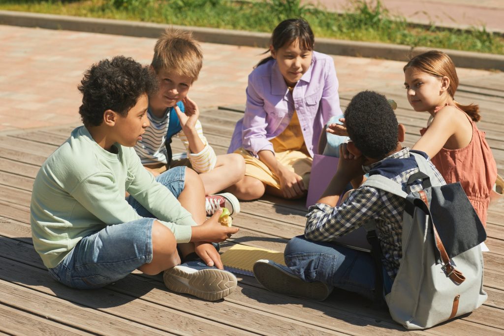 children sitting crosslegged in a circle, gathered informally outside on a wooden deck outside