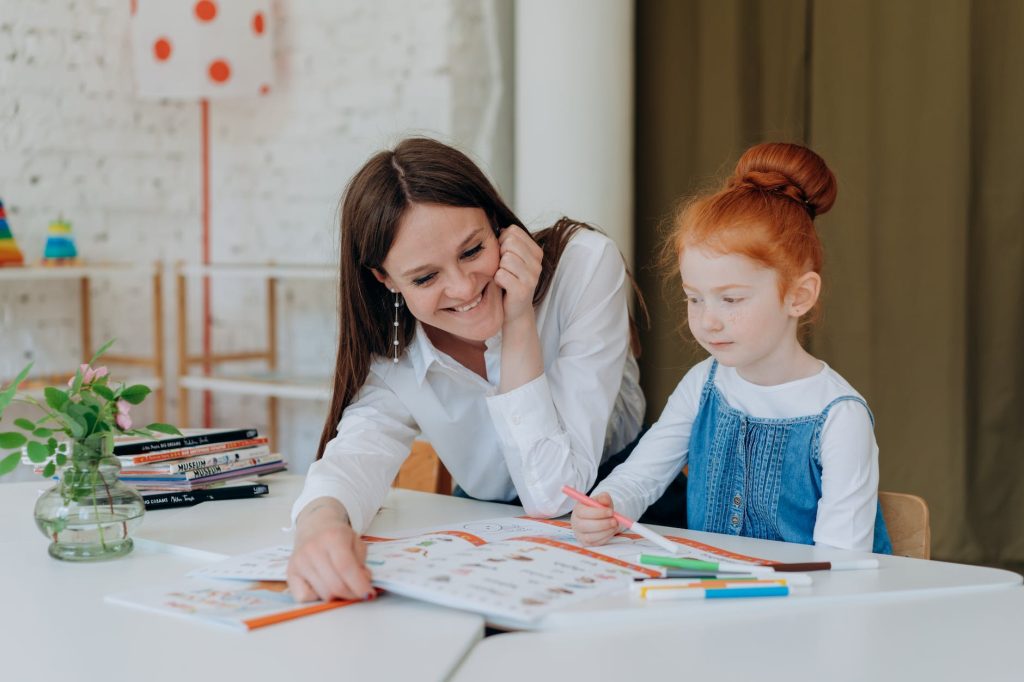 a child sitting at a table with an activity book in front of her, her mother is seated next to her, looking at the book and smiling