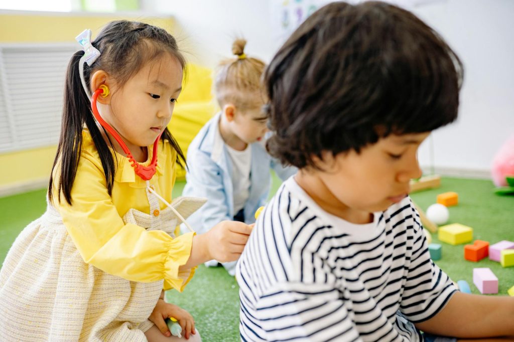 three children playing, one of them has a stethoscope and is applying the bell against the other child's back