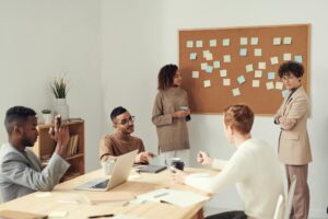 women standing beside corkboard, others are sitting at a table in a workroom