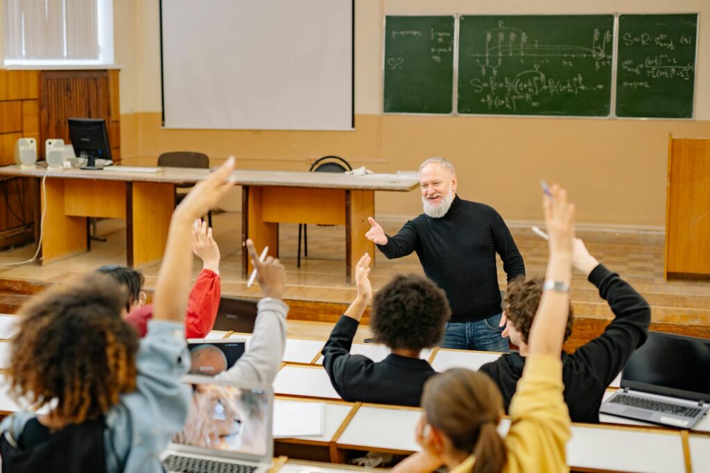 a post-secondary class with an educator in front, the students are raising their hands to answer a question